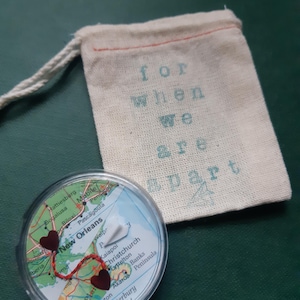 May include: A clear, round keepsake with a map design, featuring two red heart accents connected by a red thread. The map highlights New Orleans and Christchurch. A small, off-white drawstring bag with the text "for when we are apart."