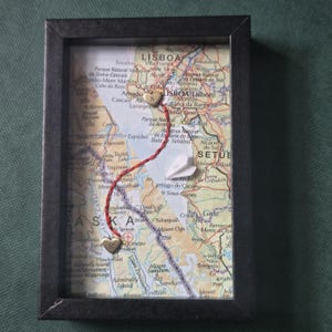 May include: A black rectangular shadow box frame containing a map of Lisbon and Alaska. A red string connects two gold-colored heart charms, one marked with the letter A and the other with the letter R. A small white paper airplane is attached to the map.