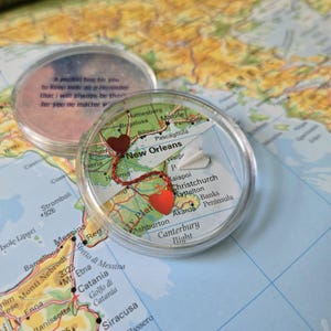 May include: Two clear, round keepsakes with map designs. One features a red string connecting two heart-shaped charms, with a paper airplane and the text "New Orleans." The other has text that reads "A pocket hug for you..." on a map background.