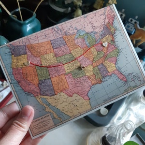 May include: A vintage map of the United States with a red line drawn across it. There are three small silver hearts pinned to the map along the red line.
