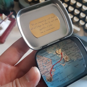 May include: A small, round tin with a map inside. The map is of the United States and Italy, with a red thread connecting two hearts. The hearts are marked with the letters "S" and "R". The tin has a gold inscription that reads: "We are connected by a red thread and will always be there for each other regardless of place time or circumstances."