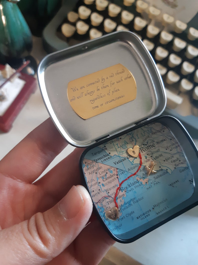 May include: A small, round tin with a map inside. The map is of the United States and Italy, with a red thread connecting two hearts. The hearts are marked with the letters "S" and "R". The tin has a gold inscription that reads: "We are connected by a red thread and will always be there for each other regardless of place time or circumstances."