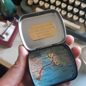 May include: A small, round tin with a map of the world inside. The map is marked with a red line and two gold hearts. The tin also has a gold inscription that reads "We are connected by a red thread and will always be there for each other regardless of place time or circumstances."