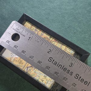 May include: A rectangular black frame containing a vintage-style map, positioned beneath a stainless steel ruler. The ruler shows the frame width is approximately 3.5 inches. The map features geographical details, text, and lines in muted tones.