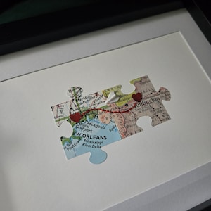 May include: A framed puzzle piece featuring a map of New Orleans and other cities, connected by a red thread and small red hearts. The puzzle piece is set against a white background.