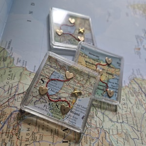May include: Three clear acrylic map coasters with gold heart and star pins connected by red thread. The coasters feature map details of various locations, including Florida and Chicago. A unique home decor item.