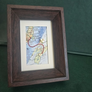 May include: A dark brown wooden frame containing a map section of Japan and Nova Scotia. A red thread connects two small gold heart charms, one marked with the letter R and the other with A, with a small gold airplane charm positioned along the path.