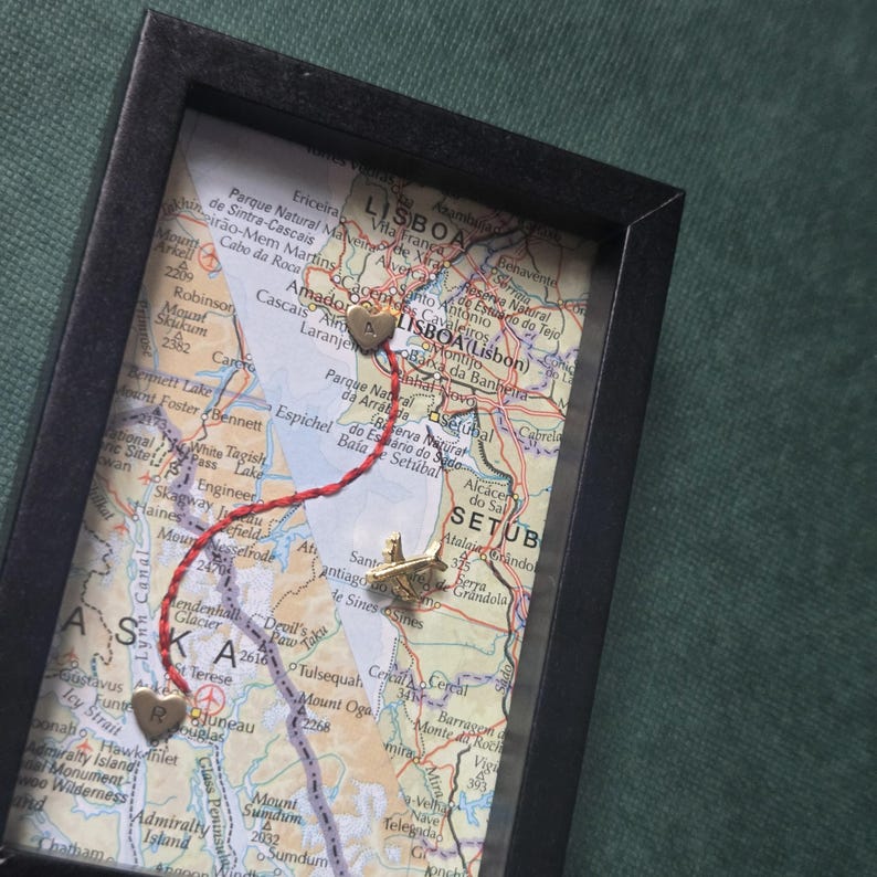 May include: A black rectangular shadow box frame containing a map of Lisbon and Alaska. A red thread connects two gold heart-shaped pins, one marked with an A and one with an R. A small gold airplane pin is positioned on the map between the two hearts.