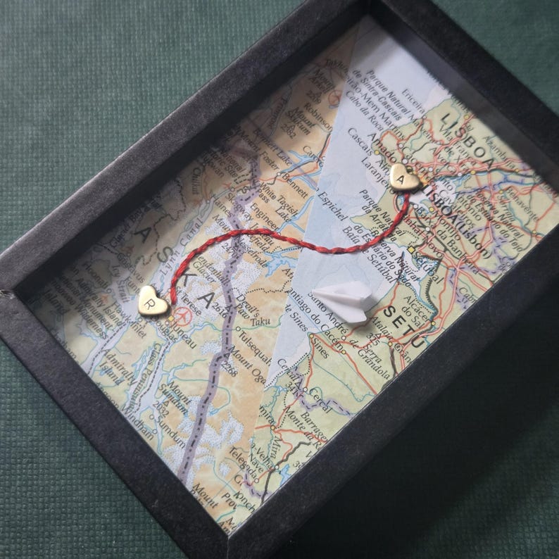 May include: A framed map display featuring a red thread connecting two small gold-colored heart charms, one engraved with the letter R and the other with the letter A. A small white paper airplane is positioned on the map surface near the letter A.