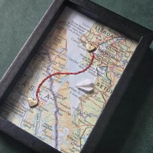 May include: A black shadow box frame containing a map collage of Alaska and Lisbon, Portugal. A red thread connects two small gold heart charms, one marked with the letter R and the other with A, alongside a small white paper airplane cutout.