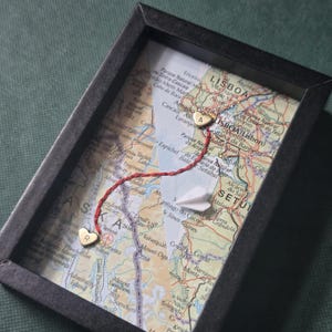 May include: A black rectangular frame containing two overlapping map sections. A red thread connects two small gold heart-shaped charms, one marked with the letter R and the other with the letter A. A small white paper airplane sits on the map surface.