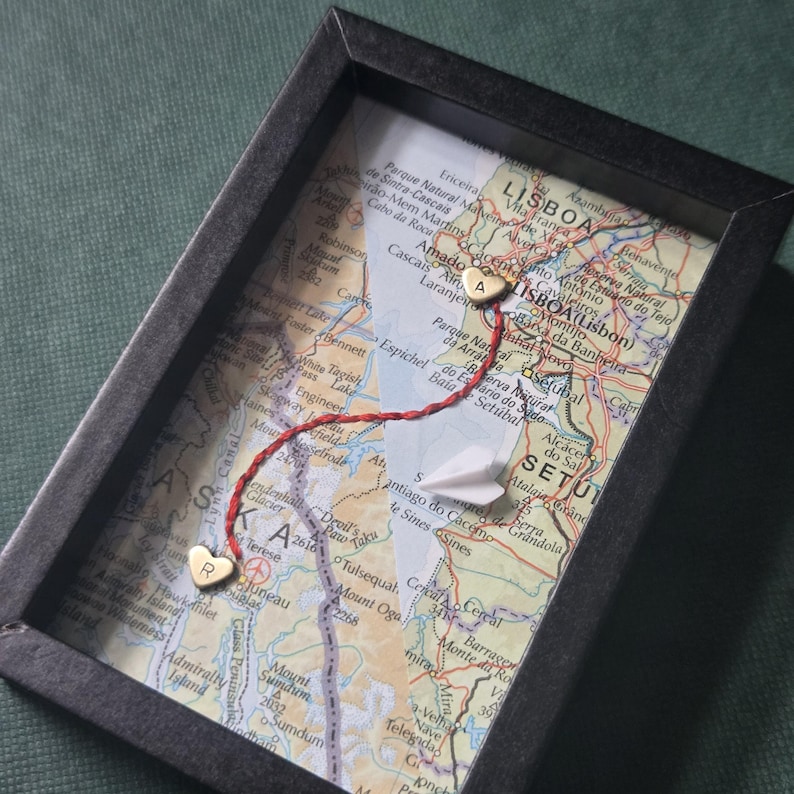 May include: A black rectangular frame containing two overlapping map sections. A red thread connects two small gold heart-shaped charms, one marked with the letter R and the other with the letter A. A small white paper airplane sits on the map surface.