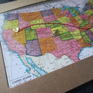 May include: A vintage map of the United States with a red thread connecting two gold heart-shaped pins. The map is framed in a brown frame and the text "UNITED STATES" is visible at the bottom.