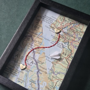 May include: A black rectangular shadow box frame containing a split map showing Alaska and Lisbon. Two gold-colored heart charms, one marked with the letter R and the other with A, are connected by a red thread. A small white paper airplane is included.