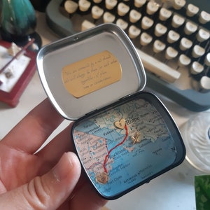 May include: A small, round tin with a map of the East Coast of the United States. The map is marked with a red line and gold hearts, with the text "We are connected by a red thread and will always be there for each other, regardless of place, time or circumstances." on the lid.