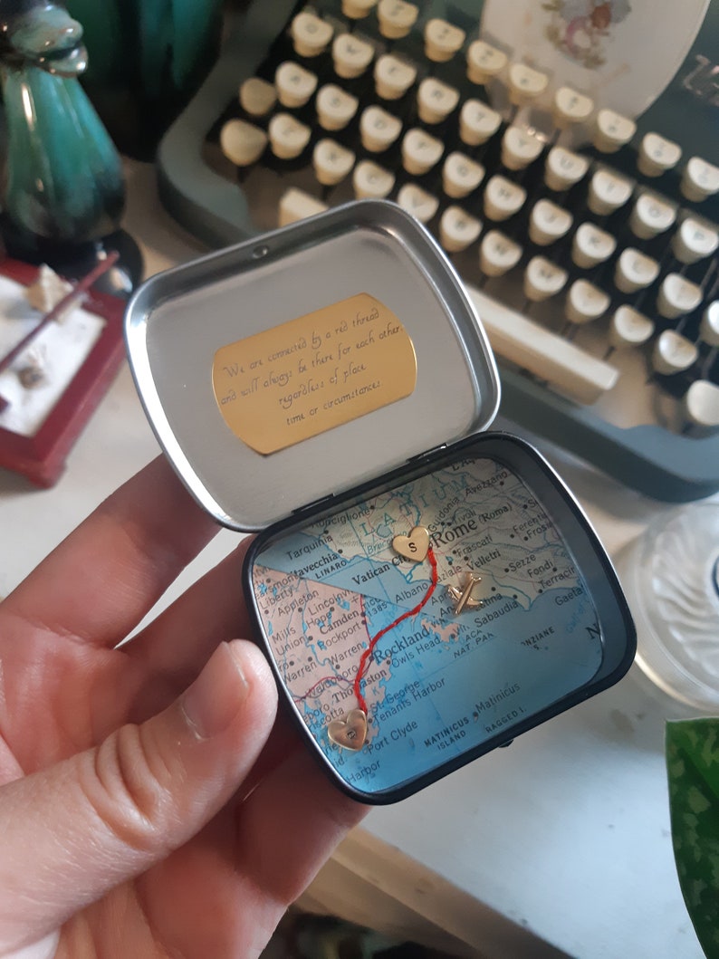May include: A small, round tin with a map of the East Coast of the United States. The map is marked with a red line and gold hearts, with the text "We are connected by a red thread and will always be there for each other, regardless of place, time or circumstances." on the lid.