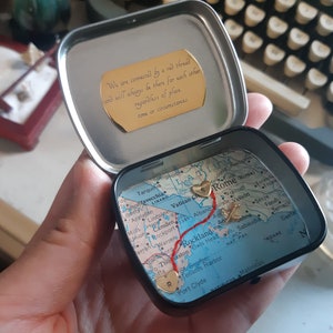 May include: A small, round tin box with a map of Italy inside. The map has a red line drawn on it, connecting two gold hearts. The tin box has a gold inscription that reads: "We are connected by a red thread and will always be there for each other regardless of place, time or circumstances."