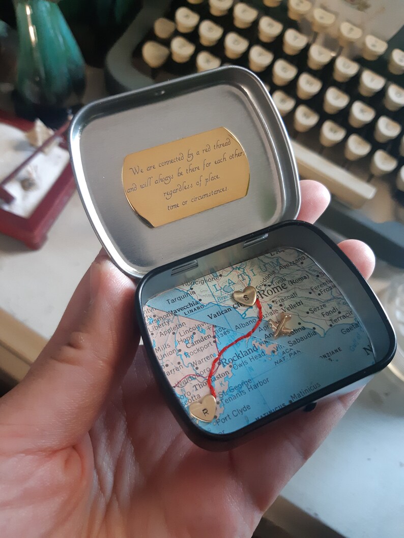 May include: A small, round tin box with a map of Italy inside. The map has a red line drawn on it, connecting two gold hearts. The tin box has a gold inscription that reads: "We are connected by a red thread and will always be there for each other regardless of place, time or circumstances."