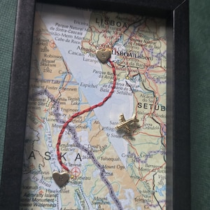 May include: A framed map display featuring a red string connecting two gold-colored heart charms, one marked with the letter A and the other with the letter R. A small gold-colored airplane charm is positioned on the map near the red string path.