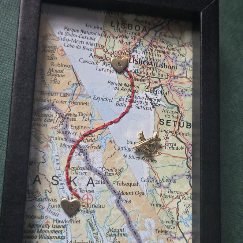 May include: A framed map display featuring a red string connecting two gold-colored heart charms, one marked with the letter A and the other with the letter R. A small gold-colored airplane charm is positioned on the map near the red string path.