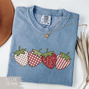 May include: A blue jean colored t-shirt with four strawberry appliqués. Each strawberry has a different pattern: white polka dots, red gingham, red floral, and red and white stripes. A gold necklace with a pendant rests on the shirt.