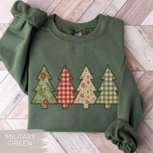 May include: Military green sweatshirt with four Christmas tree appliqués. The trees feature floral and plaid patterns in red, green, and white. The sweatshirt has a crew neck and long sleeves.