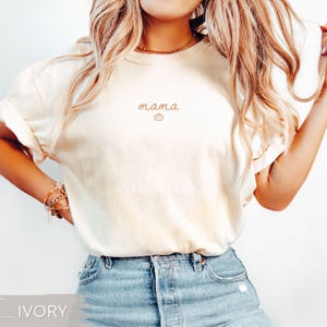 May include: Ivory-coloured t-shirt featuring the word "mama" and a pumpkin graphic in light brown. The shirt has rolled-up sleeves and is worn with blue jeans. The word "IVORY" is at the bottom of the image.