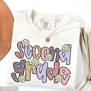 May include: Ivory-colored t-shirt with the words "Second Grade" in a colorful, floral design. The text is accented with daisy graphics. A gold necklace is draped across the shirt.