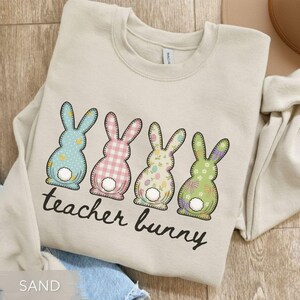 May include: Sand-colored sweatshirt featuring four colorful Easter bunny designs. Each bunny has a unique pattern, including polka dots, gingham, and floral prints. The text "teacher bunny" is printed below the bunnies in a script font.