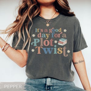 May include: Dark gray t-shirt with the text "It's a good day for a Plot Twist" in colorful letters. The shirt features a stack of books graphic and small flower accents. The word "PEPPER" is at the bottom.
