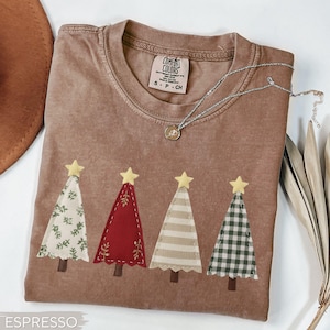 May include: Espresso-colored t-shirt with four appliqued Christmas trees. Each tree has a different pattern: floral, red, striped, and plaid. The trees are topped with yellow stars. A silver necklace with a pendant is draped across the shirt.