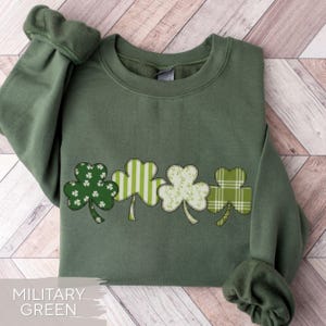 May include: Military green sweatshirt featuring a row of four shamrock designs. Each shamrock has a different pattern, including white floral, stripes, and plaid. The sweatshirt is made of a soft material and is perfect for St. Patrick's Day.