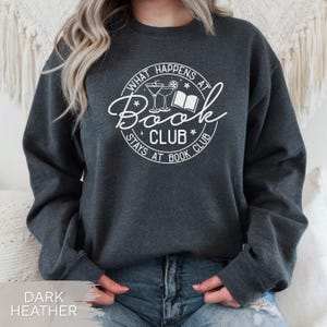 May include: Dark heather gray sweatshirt with a white graphic. The graphic reads "What Happens at Book Club Stays at Book Club" in a circular design with a book and cocktails. The sweatshirt is a casual style.
