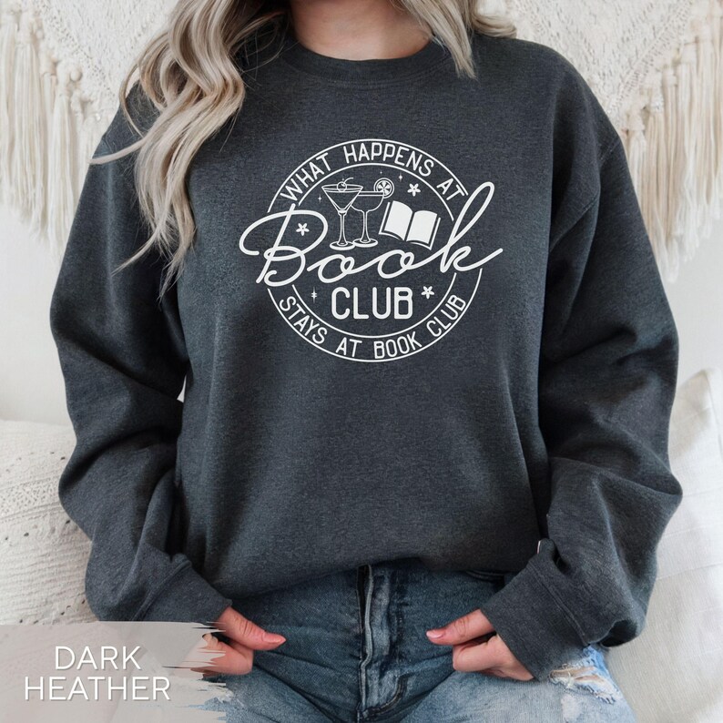 May include: Dark heather gray sweatshirt with a white graphic. The graphic reads "What Happens at Book Club Stays at Book Club" in a circular design with a book and cocktails. The sweatshirt is a casual style.