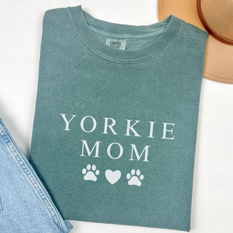 May include: A teal blue t-shirt with the text "Yorkie Mom" in white lettering and three white paw prints and a heart.