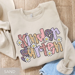 May include: Sand-colored sweatshirt with the word "Kindergarten" in a colorful, floral design. The letters are decorated with patterns and daisy accents. The sweatshirt is folded, with a portion of a pair of jeans visible.