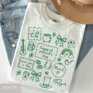 May include: White t-shirt with green St. Patrick's Day themed illustrations, including a pot of gold, shamrocks, and a rainbow. The text "happy st. patrick's day" is also featured. The shirt is folded on top of blue jeans.