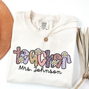 May include: Cream-colored t-shirt with the word "teacher" in a colorful floral design, with the name "Mrs. Johnson" below. A gold necklace with a pendant and a brown hat are also visible.
