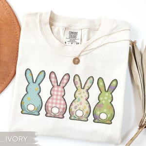 May include: Ivory-colored t-shirt featuring four colorful Easter bunny designs. Each bunny has a unique pattern: blue with yellow stars, pink gingham, floral, and green with bows. A gold necklace with a pendant rests on the shirt.