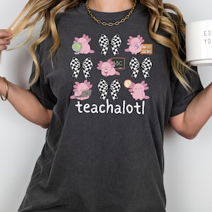 May include: Dark grey t-shirt with a design of pink axolotls, chequered bows, and the text "teachalot!". The axolotls are shown with school-related items. A gold chain necklace is visible.