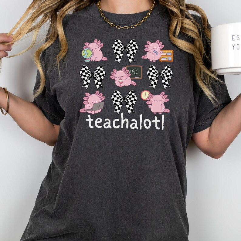 May include: Dark grey t-shirt with a design of pink axolotls, chequered bows, and the text "teachalot!". The axolotls are shown with school-related items. A gold chain necklace is visible.