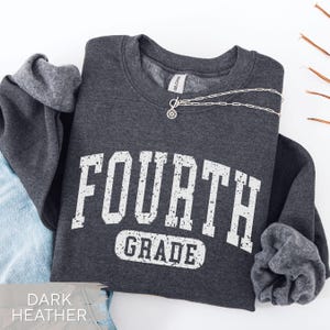 May include: A dark heather gray crewneck sweatshirt featuring the white distressed text "FOURTH GRADE" in a collegiate font. A silver-toned chain necklace with a small circular pendant rests on the neckline of the garment.