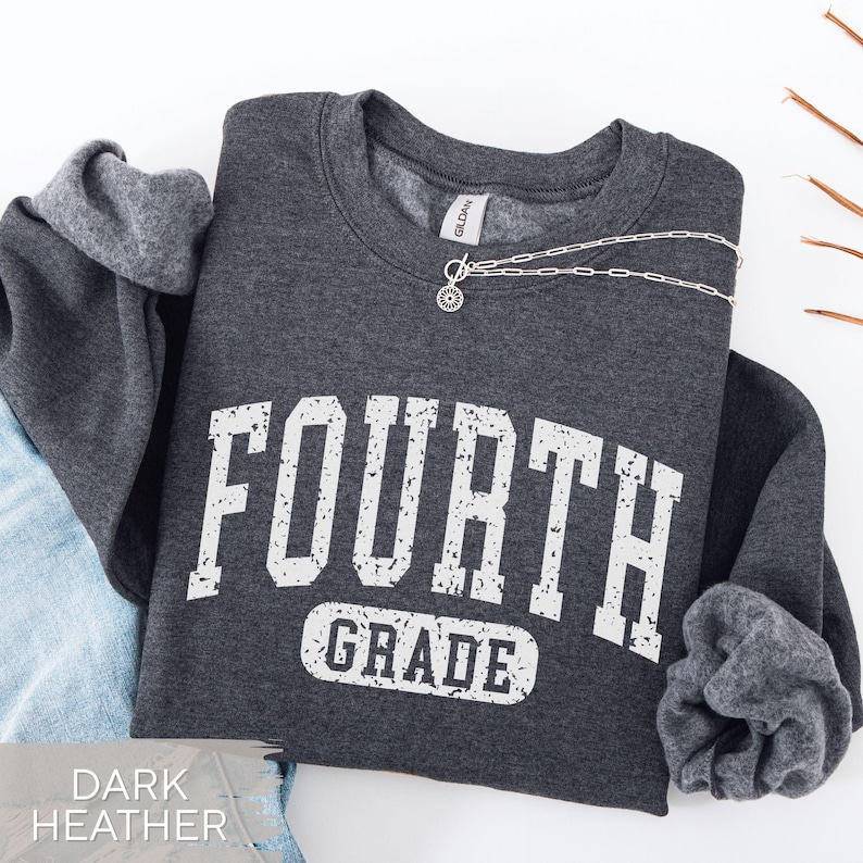 May include: A dark heather gray crewneck sweatshirt featuring the white distressed text "FOURTH GRADE" in a collegiate font. A silver-toned chain necklace with a small circular pendant rests on the neckline of the garment.