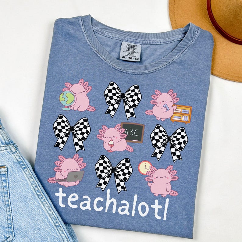 May include: A blue Comfort Colors t-shirt with a design of pink axolotls with school-themed accessories and black and white checkered bows. The word "teachalotl" is printed in white at the bottom of the shirt.