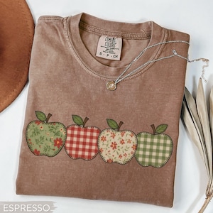 May include: Espresso-colored t-shirt featuring four apple designs with various patterns: floral, gingham, and solid. The shirt has a crew neck and short sleeves. A silver necklace with a pendant is draped across the shirt. The tag reads "Comfort Colors".