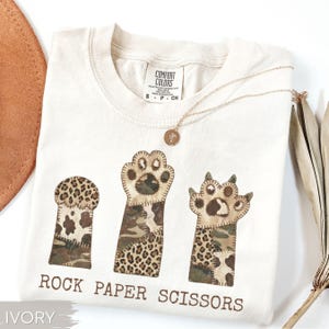 May include: Ivory-colored t-shirt with a "Rock Paper Scissors" design. The design features three paw-shaped patches with leopard and camouflage patterns. A gold necklace with a pendant rests on the shirt.