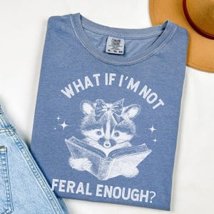 May include: A blue t-shirt with a white graphic of a raccoon reading a book. The text on the shirt reads "What if I'm not feral enough?"