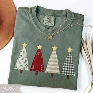 May include: A moss green t-shirt featuring four Christmas tree appliqués with yellow star toppers. The trees have different patterns: floral, red with leaf accents, striped, and plaid. A gold necklace with a pendant is draped across the shirt.