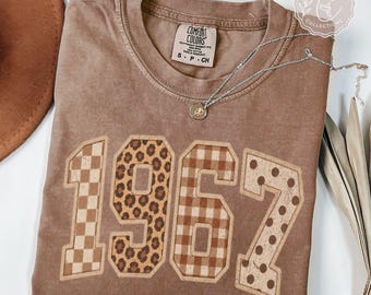 May include: A brown short-sleeve t-shirt featuring the number 1967 in large block letters with varied patterns including checkerboard, leopard print, gingham, and polka dots. A silver chain necklace with a small pendant rests on the shirt.