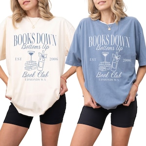 May include: Two t-shirts, one cream and one blue, each with the text "BOOKS DOWN Bottoms Up Book Club EDMONDS WA EST 2006" in a navy blue design. The shirts are short-sleeved and worn with black shorts.